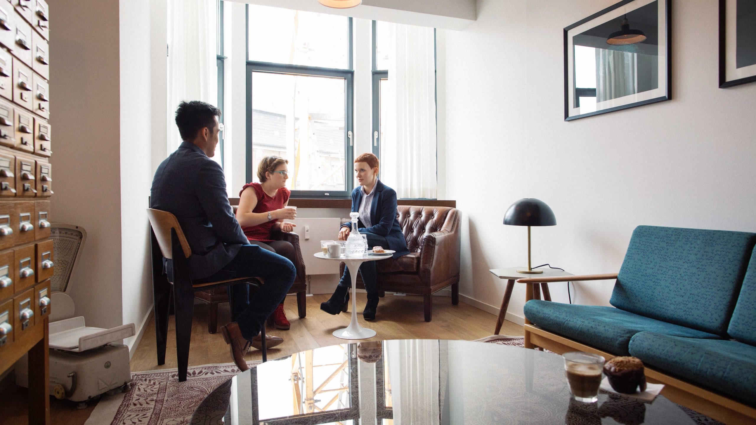 Three Young People Chatting Over Coffee In Bright Minimalist Design Lounge